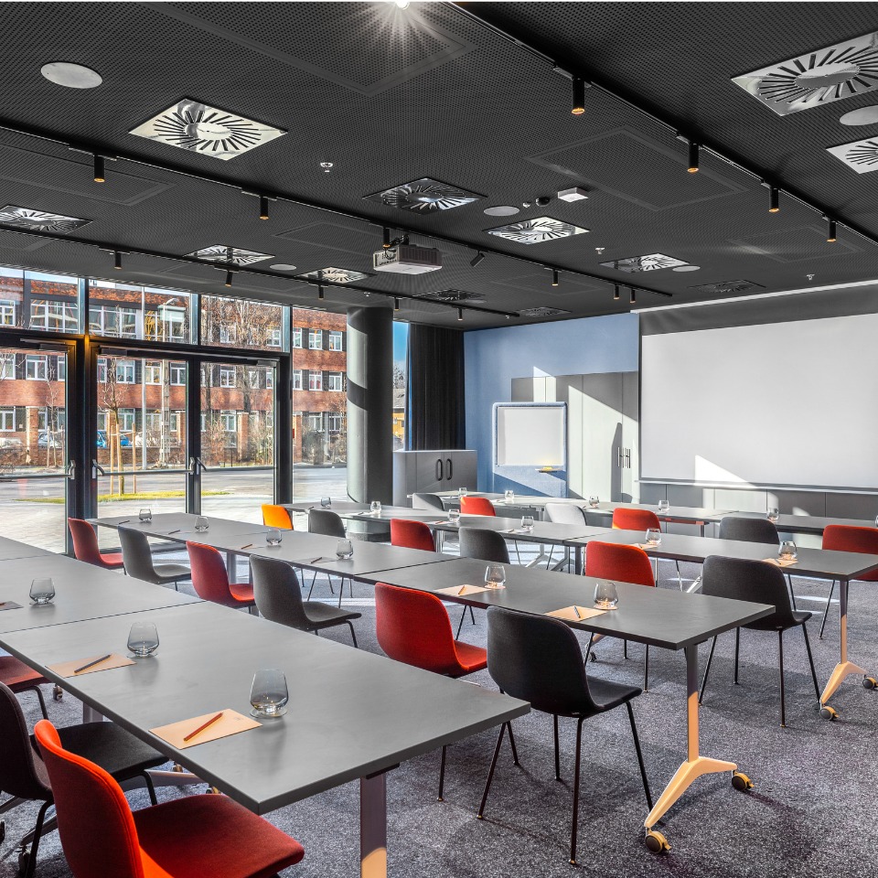 Conference room with grey tables, colorful chairs, and a large screen, featuring natural light from windows.