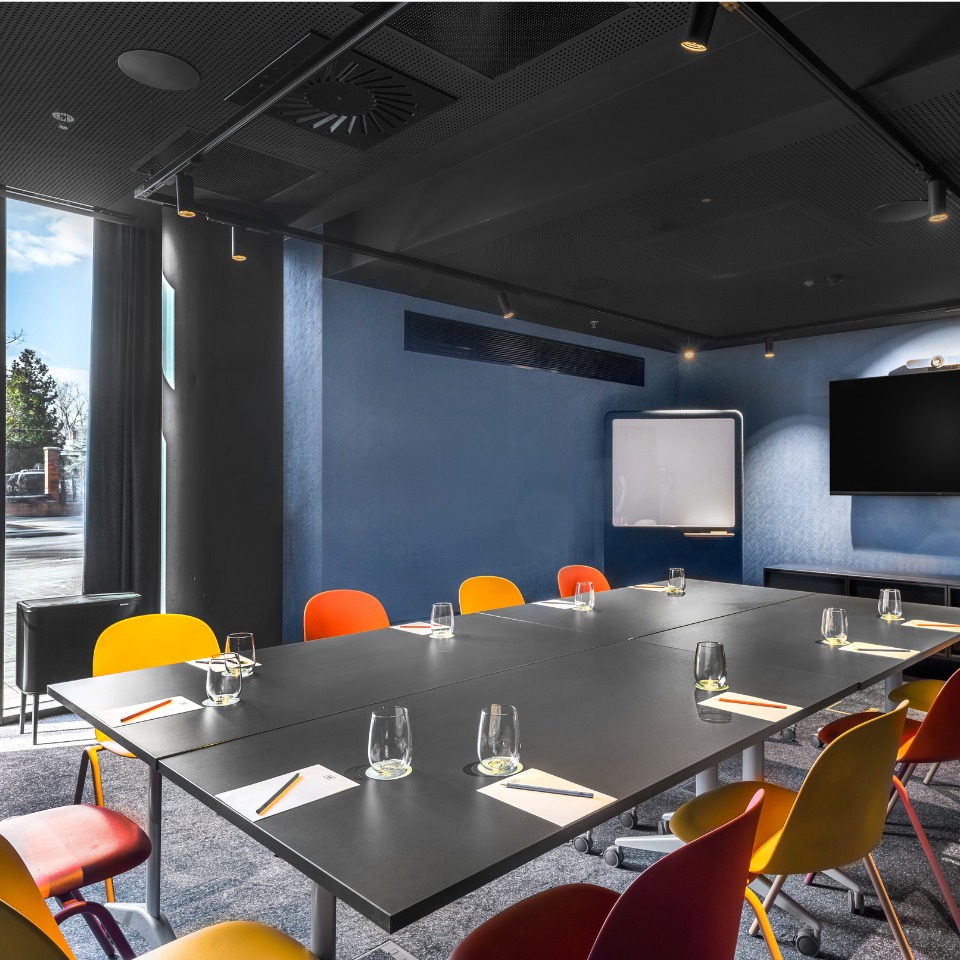 A conference room featuring a long table, colorful chairs, glass water cups, a whiteboard, and a TV screen.