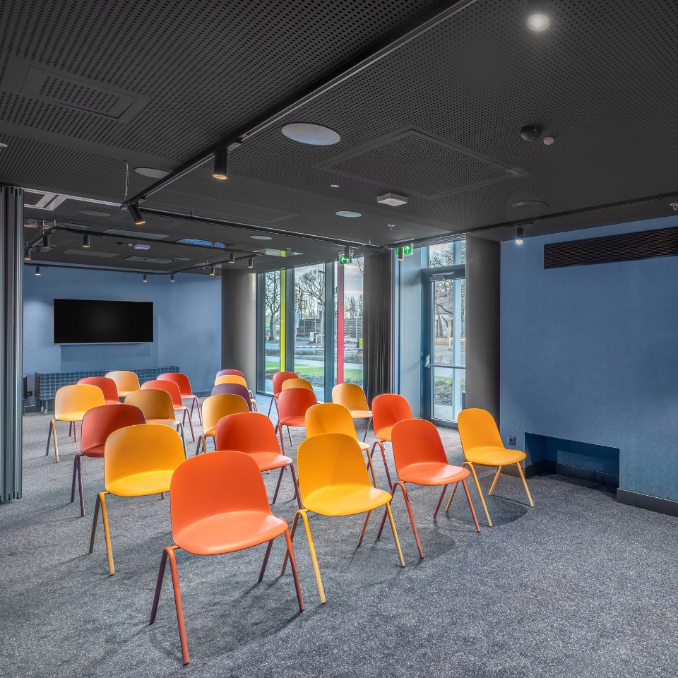 A conference room featuring bright orange and yellow chairs arranged in rows, with large windows and a wall-mounted TV.