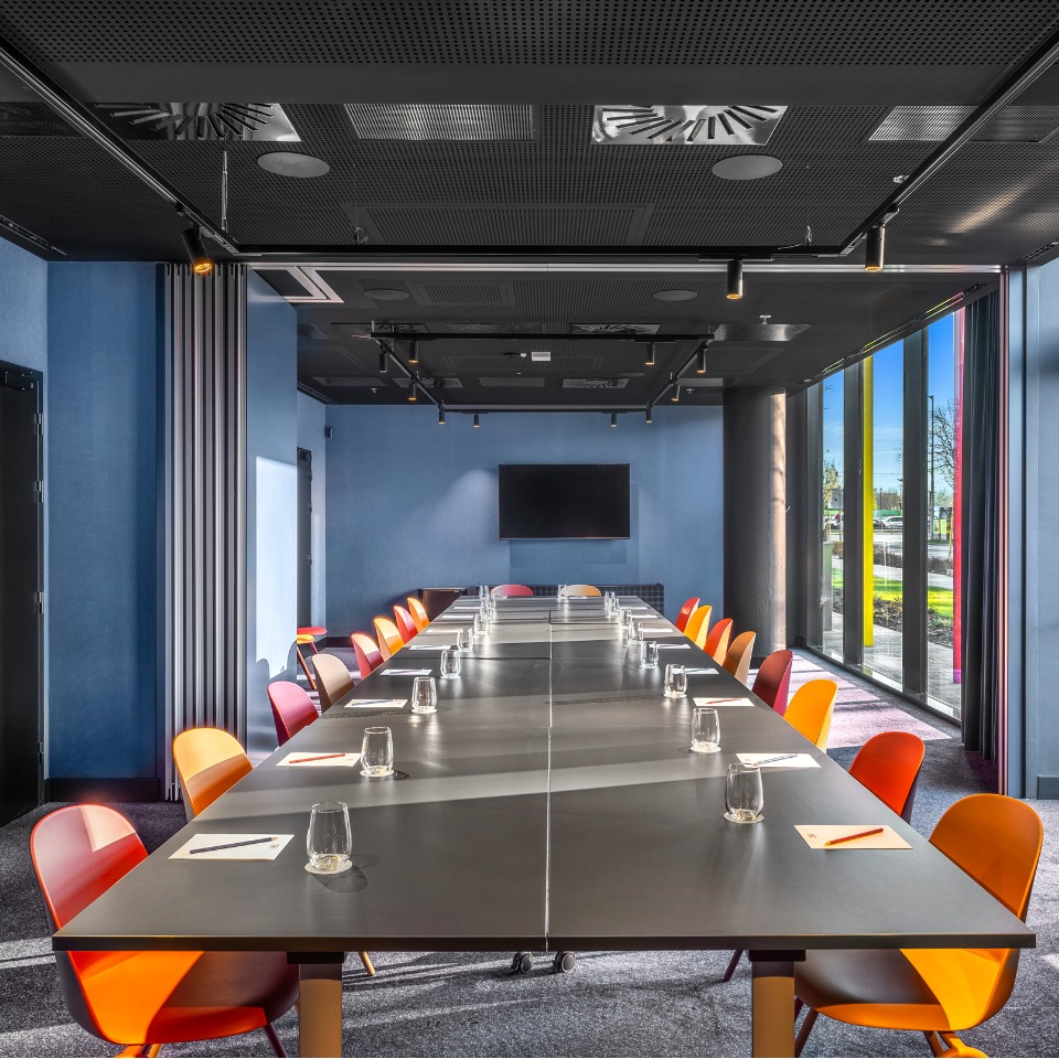 Conference room with a long table, colorful chairs, and a large screen, filled with natural light from floor-to-ceiling windows.