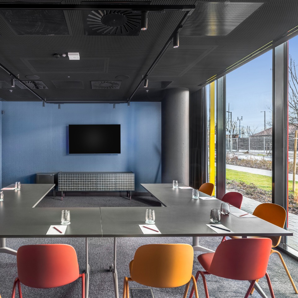 Conference room with a dark ceiling, blue walls, a TV screen, and colorful chairs around a sleek table, featuring large windows.