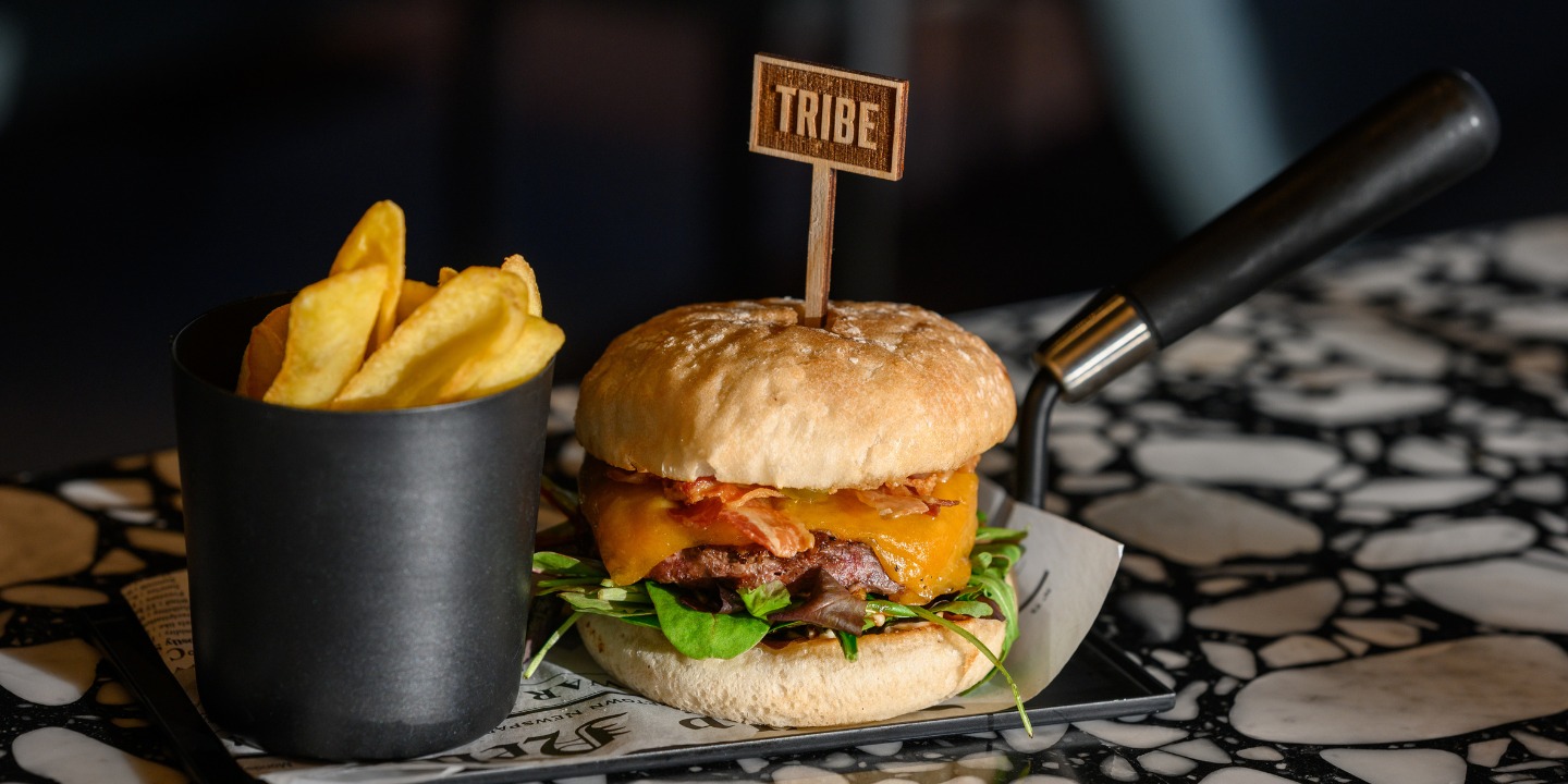 A gourmet burger with cheddar, bacon, and greens, topped with a "TRIBE" marker, accompanied by a side of golden fries.