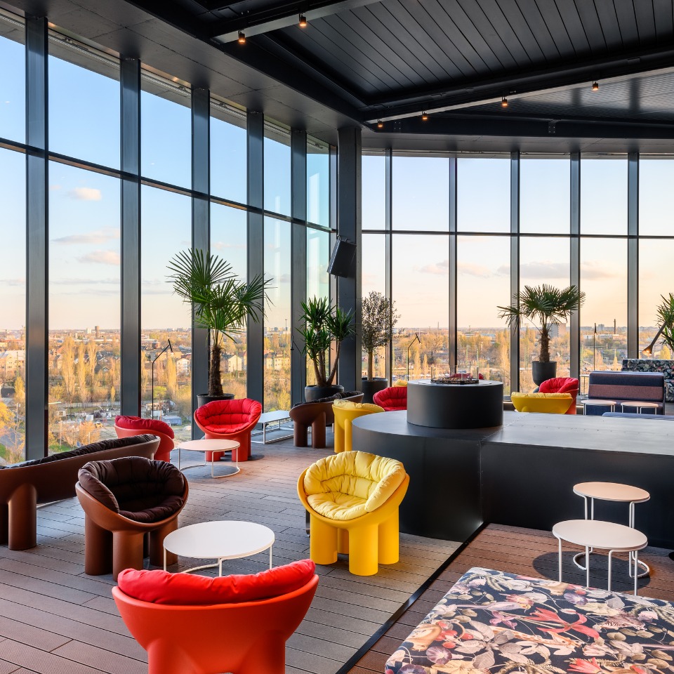 A stylish var with colorful seating, large windows, and a panoramic city view, adorned with plants and modern decor.