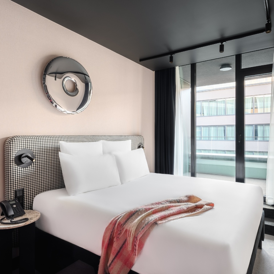 Hotel room featuring a stylish bed, decorative mirror, and balcony doors with sheer curtains, accented by a cozy blanket.