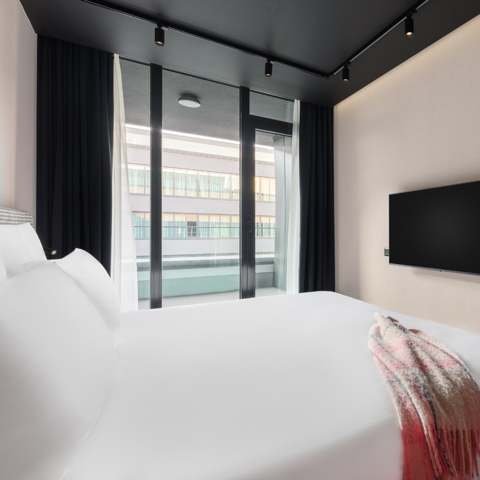 Hotel room with a king-sized bed, sleek decor, and large windows overlooking a balcony area. TV mounted on the wall.