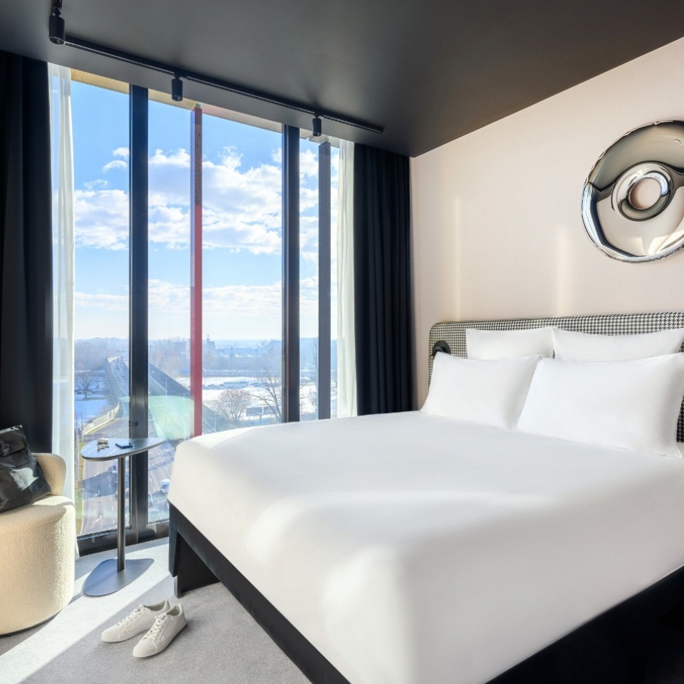 Hotel room featuring a large bed, stylish furniture, and floor-to-ceiling windows with scenic views and bright daylight.