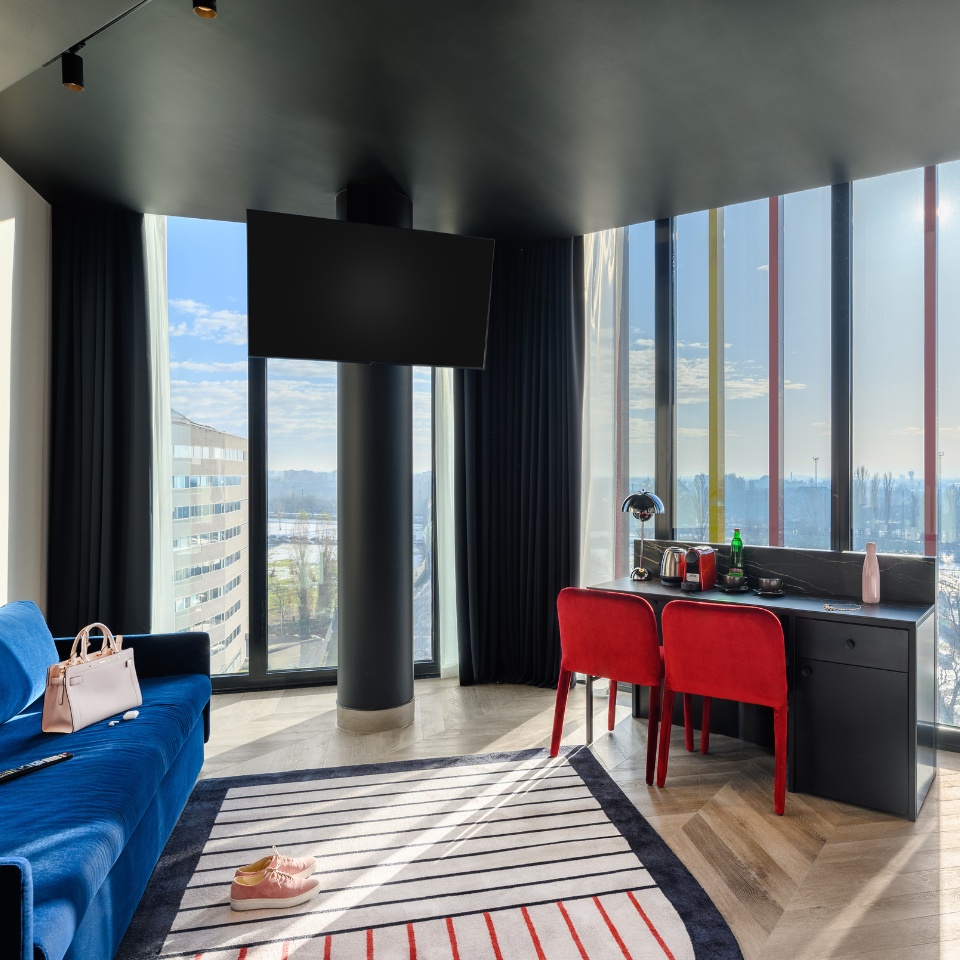A stylish hotel room featuring a blue sofa, red chairs, and a desk by large windows, with a view of the city skyline.