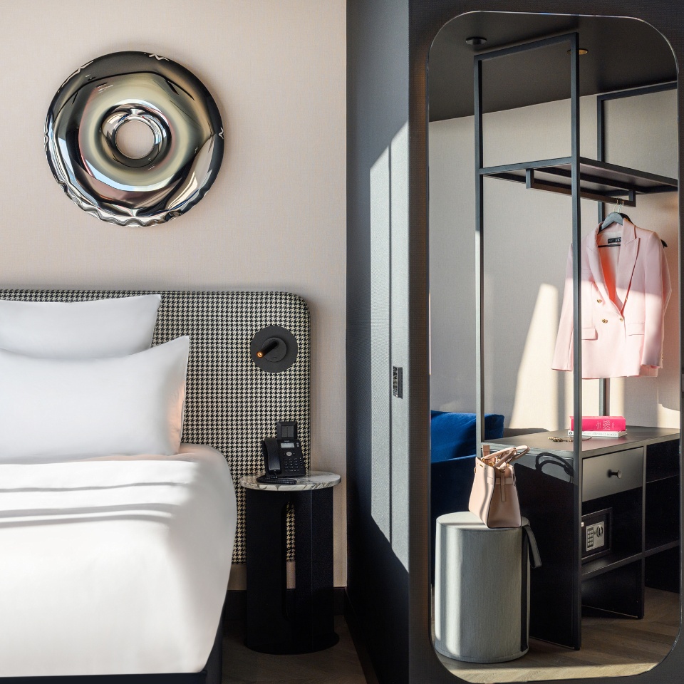 A modern hotel room featuring a stylish bed with white pillows, a sleek nightstand, and a minimalist clothing area with a pink blazer.