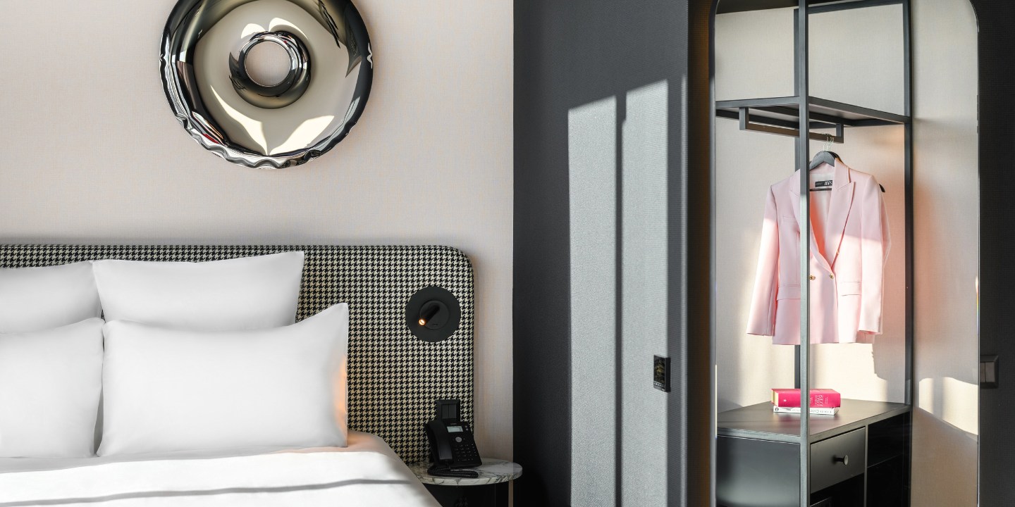 A stylish hotel room featuring a plush bed with white pillows, a sleek pink blazer hanging in a modern closet, and a bedside phone.