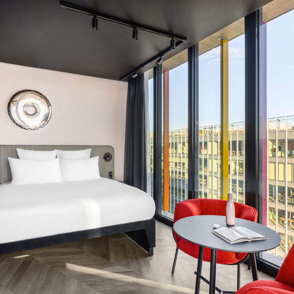 A hotel room featuring a king bed, vibrant red chairs, and large windows with colorful frames and natural light.