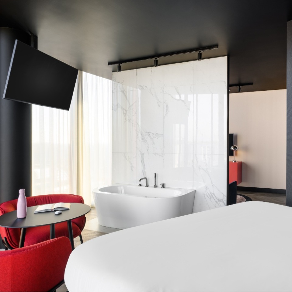 Hotel room featuring a stylish soaking tub, sleek furnishings, and large windows with flowing curtains. Cozy seating and TV included.