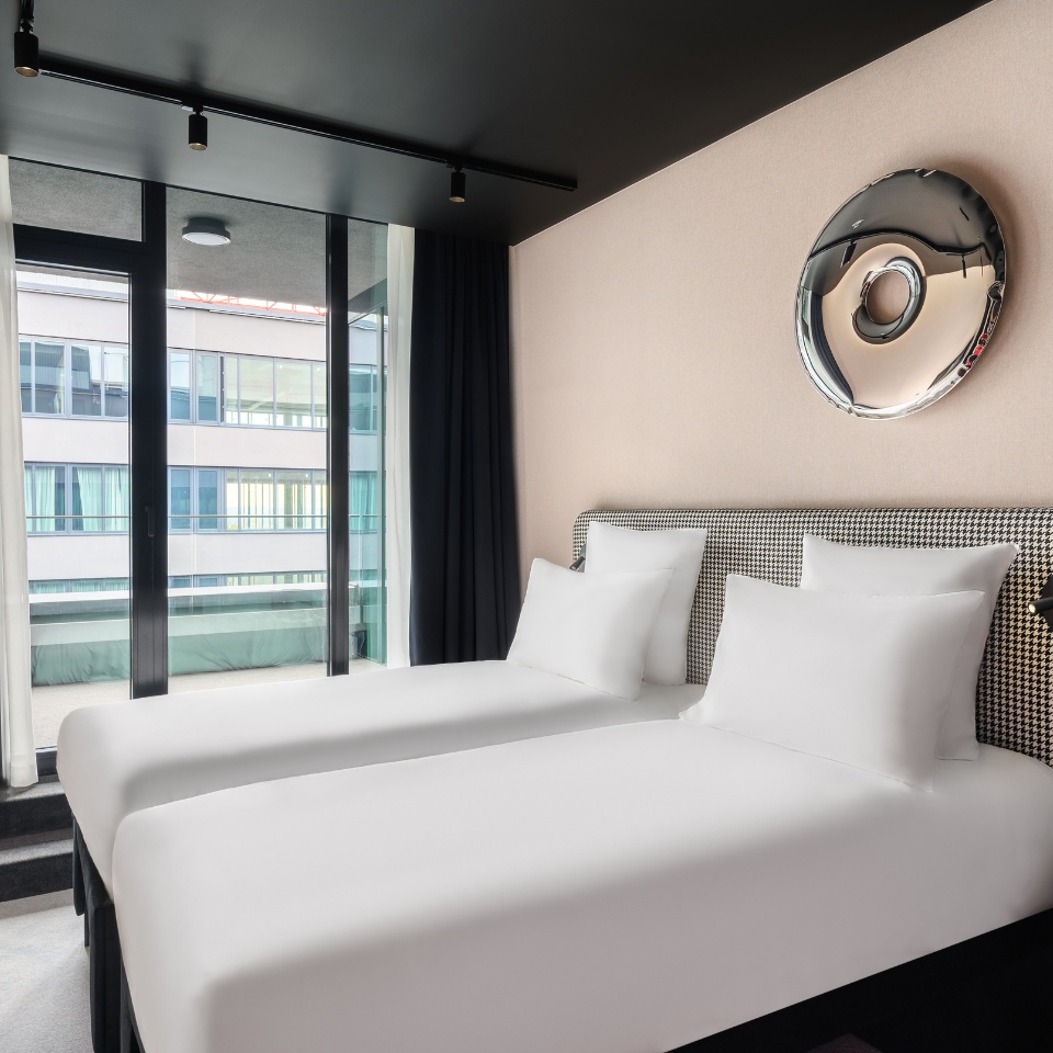 Hotel room featuring two neatly made twin beds, large windows with natural light, and stylish decor.