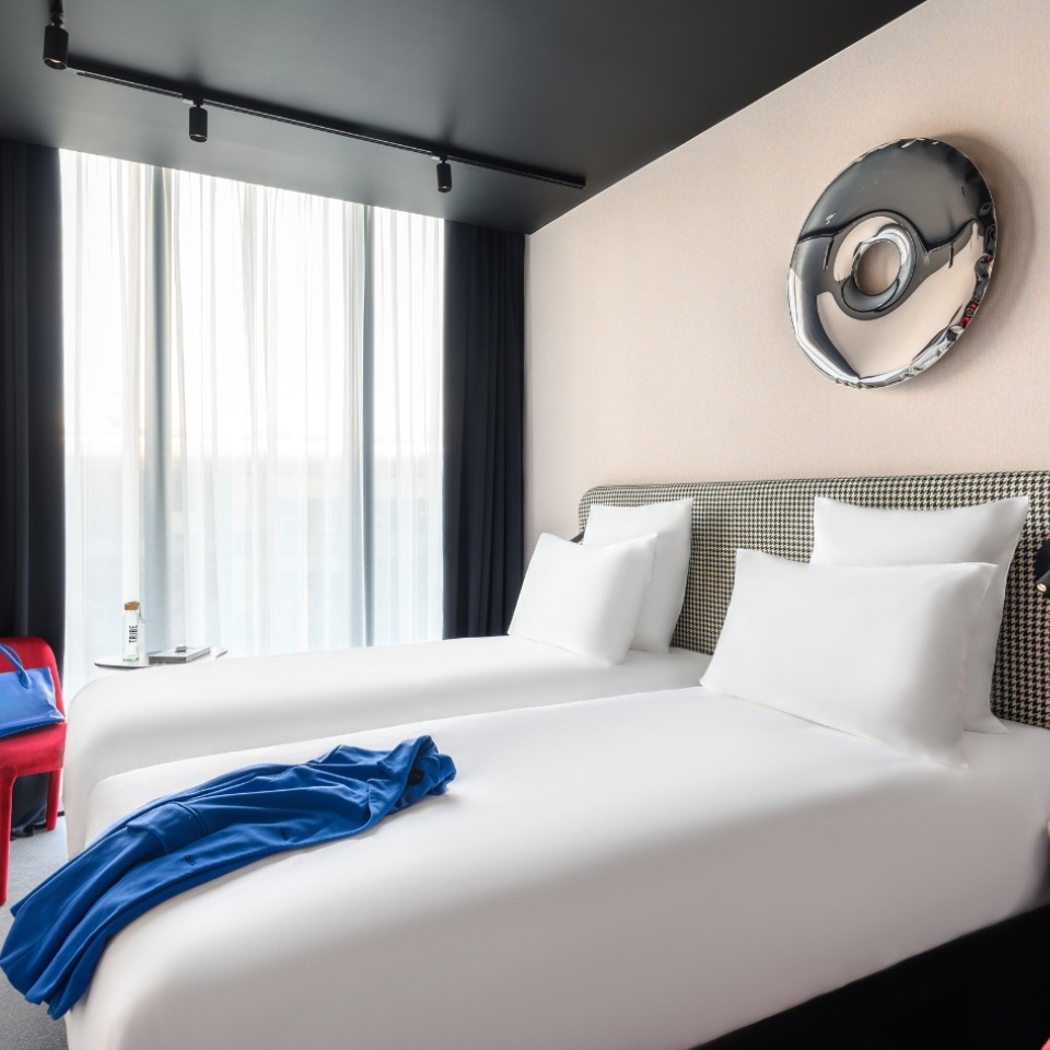 Hotel room featuring two neatly made beds, a blue garment draped on one, and artistic metal decor, with light filtering through sheer curtains.