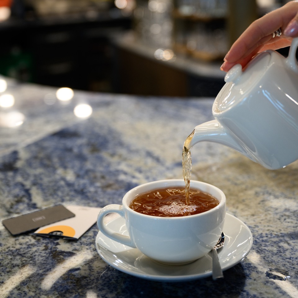 A hand pours hot tea from a white teapot into a matching cup on a marble countertop. The scene conveys warmth and relaxation.