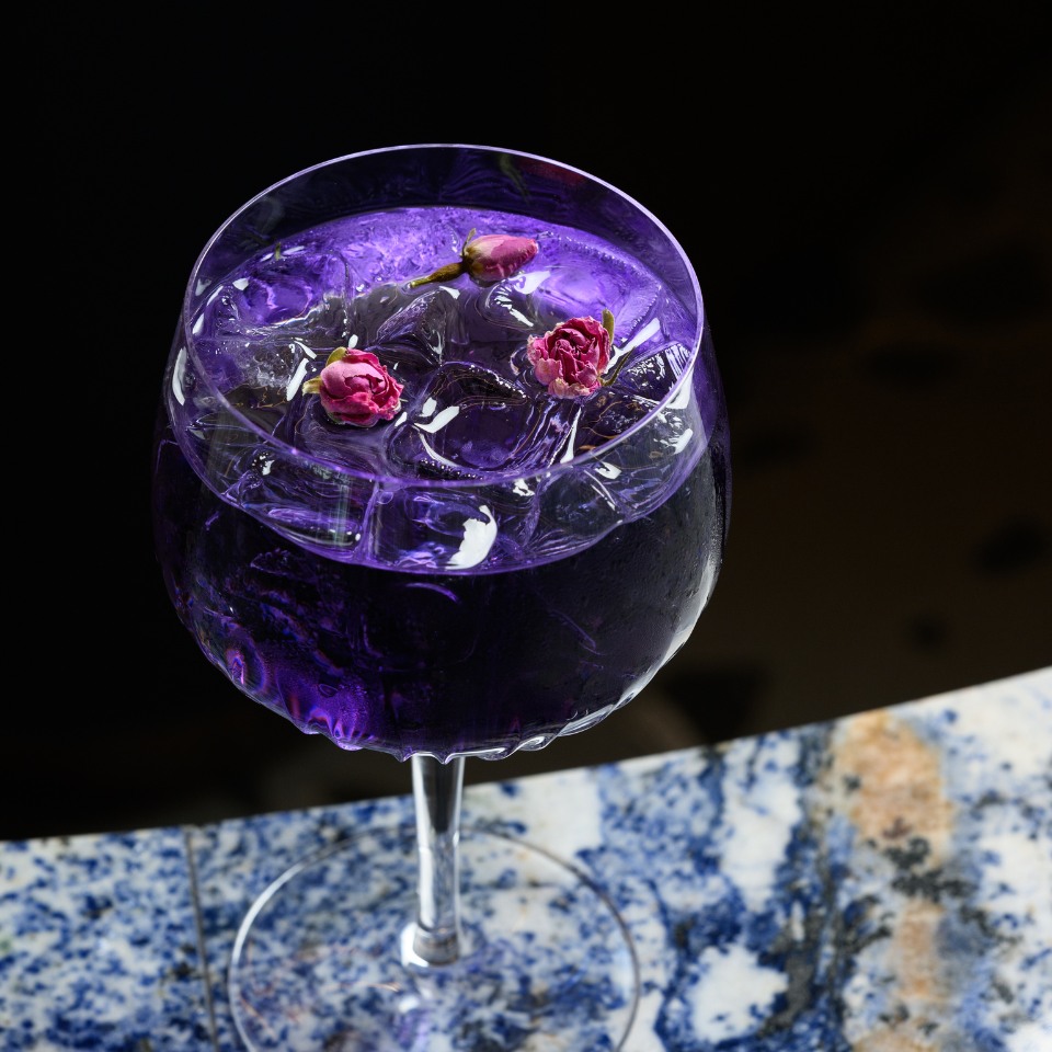A vibrant purple cocktail in a glass with ice cubes and pink rosebuds, set against a blue marble table. The mood is elegant and luxurious.