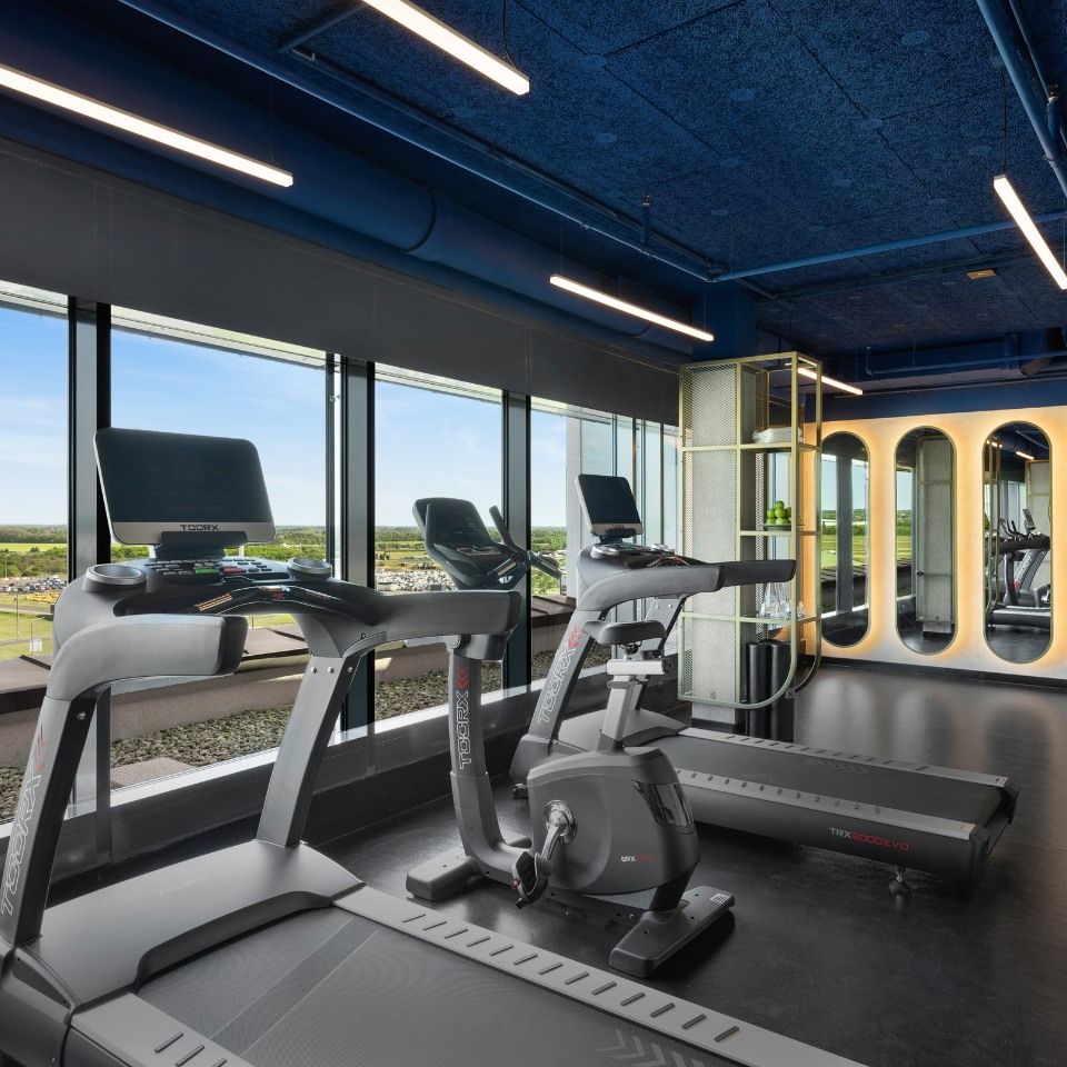 Modern gym with large windows showcasing a scenic view, featuring treadmills and an exercise bike. Sleek design, bright lighting, and mirrored wall.