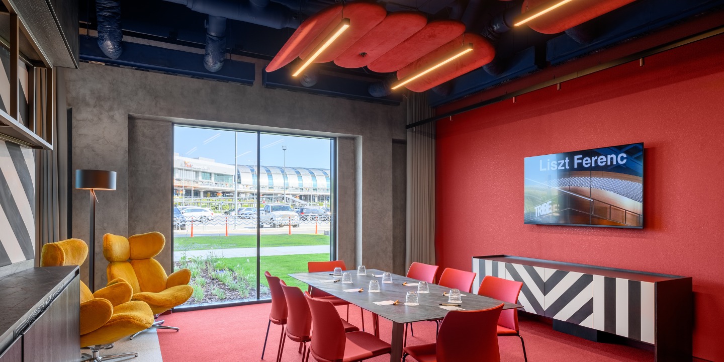 Modern meeting room with red walls and carpet, featuring a large table surrounded by red chairs. A cozy yellow armchair sits near a large window with a view outside. The ceiling has red acoustic panels with integrated lights, creating a stylish, vibrant atmosphere.