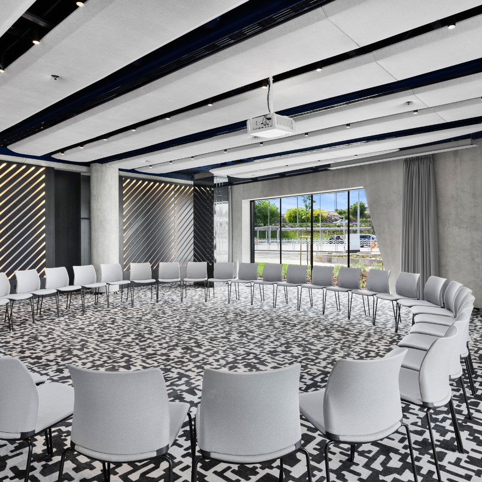 Modern meeting room with a large circular arrangement of white chairs on a patterned carpet. Diagonal wall patterns and large windows provide a bright, open feel.