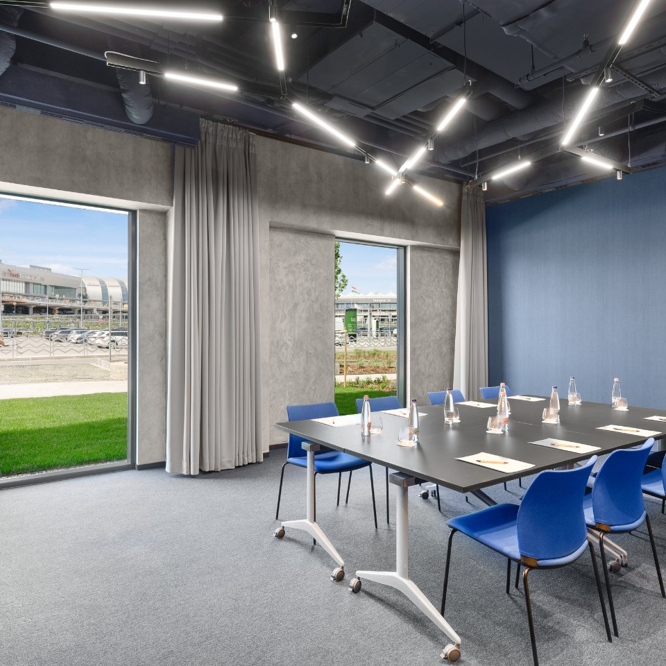 Modern conference room with a sleek black table, blue chairs, and bottled water. Large windows offer a view of a building and green lawn outside.