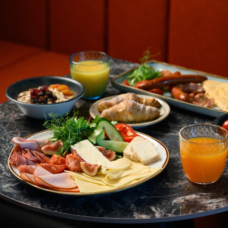 A diverse breakfast spread on a table includes cold cuts, cheeses, figs, and cucumbers, with croissants, oatmeal, and orange juice in the background.