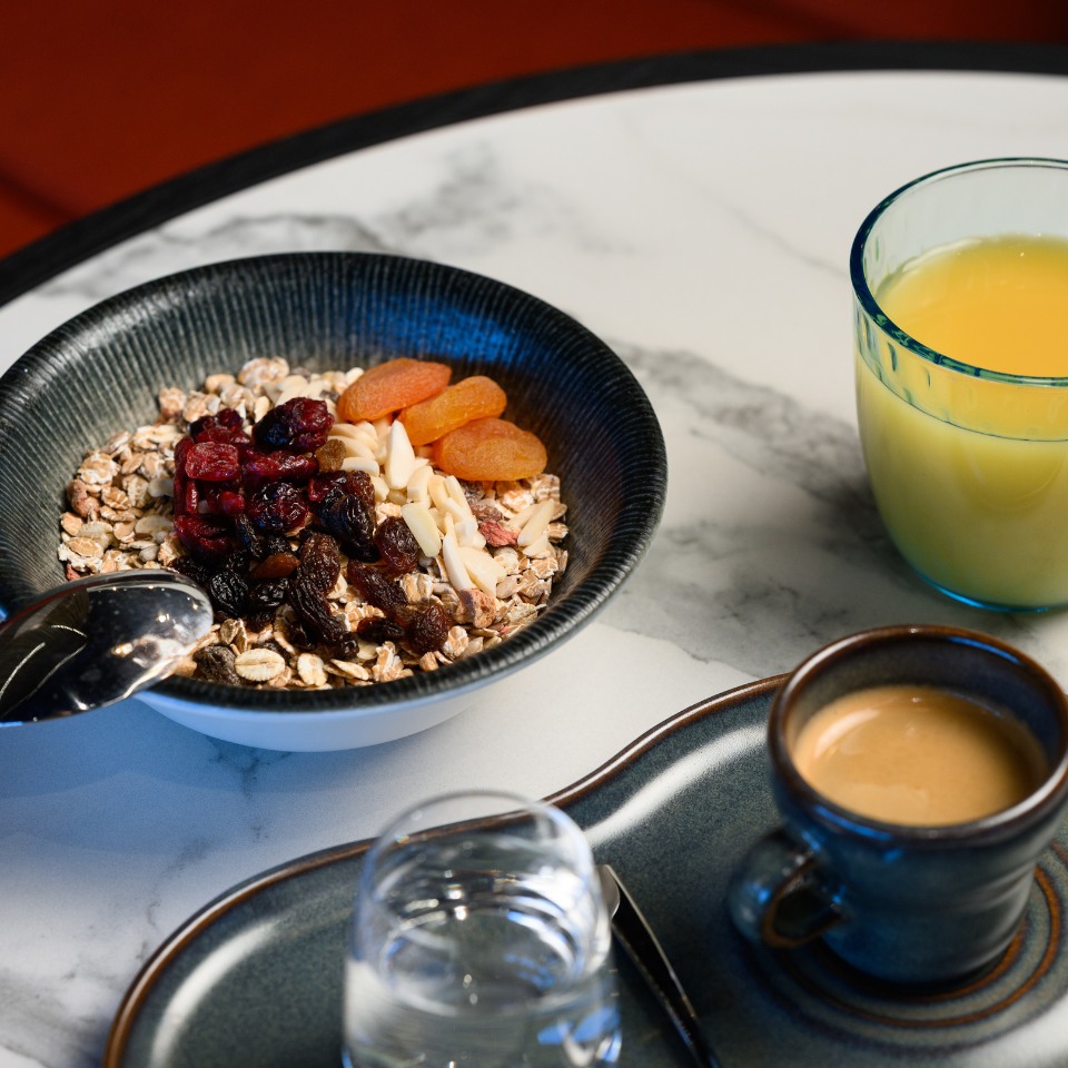 A bowl of muesli topped with dried fruits and nuts, alongside a glass of orange juice and a cup of espresso on a marble table. The scene is calm and inviting.