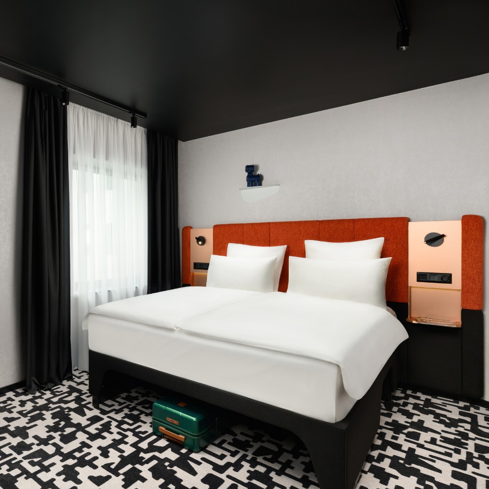 Modern hotel room with a double bed featuring crisp white linens, orange headboard, and geometric black-and-white carpet. Soft lighting and dark curtains.