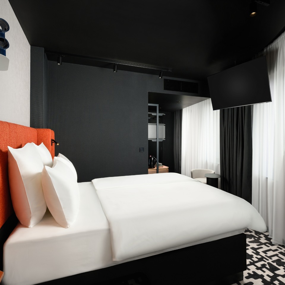 A modern hotel room with a sleek black and white color scheme. It features a neatly made bed with white linens, and an orange headboard. Black walls and curtains contrast with the room's bright light, creating a stylish atmosphere. A wall-mounted TV and a small seating area are visible.