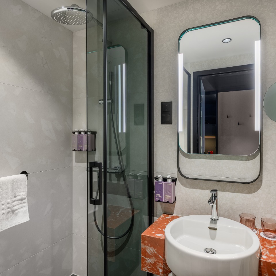 Modern bathroom with a sleek shower enclosure, glass doors, and a rainfall showerhead. A round white sink sits on an orange marble counter with a mirror above.