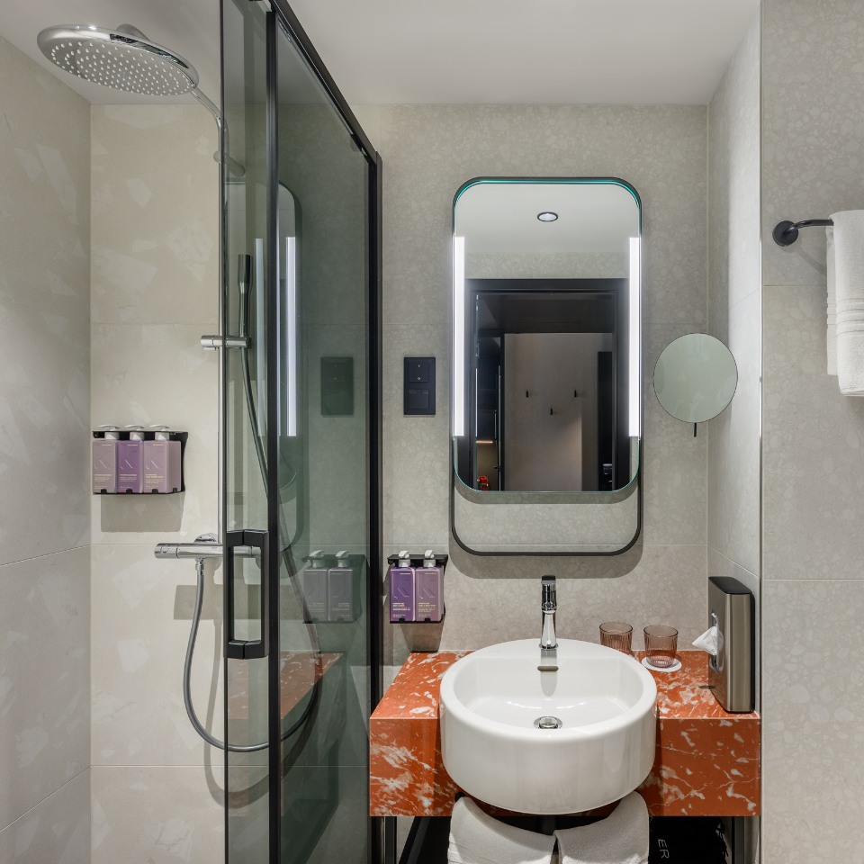 Modern bathroom with glass shower, overhead rain shower head, and soap dispensers. A round sink on a red marble counter, illuminated mirror.