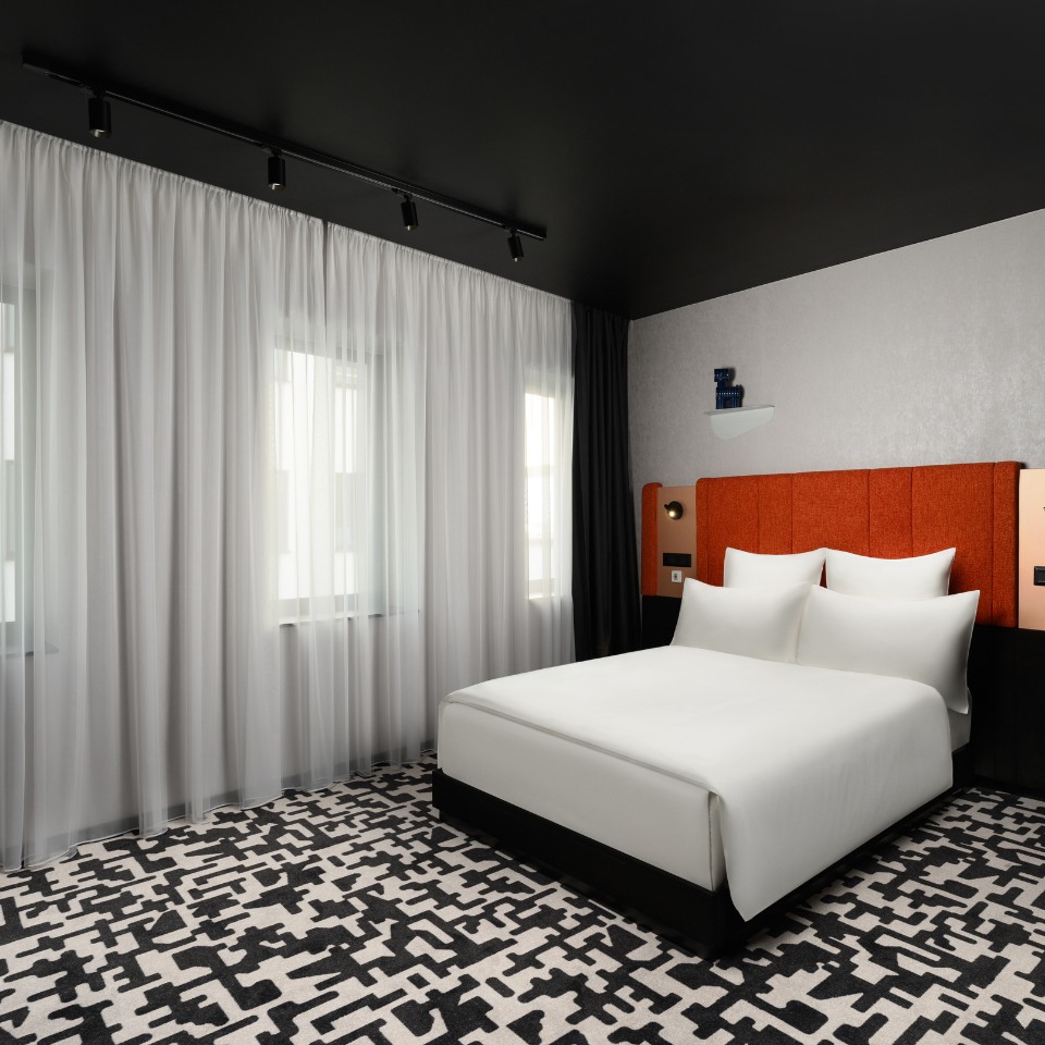 Modern bedroom with clean lines features a white bed, orange headboard, abstract-patterned carpet, sheer curtains, and a black ceiling, creating a sleek, serene ambiance.