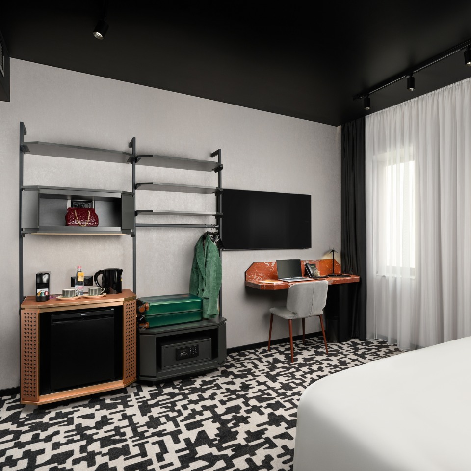 Modern hotel room with geometric black-and-white carpet, open shelving, TV, desk with a laptop, and a window with sheer curtains. Cozy and sleek.