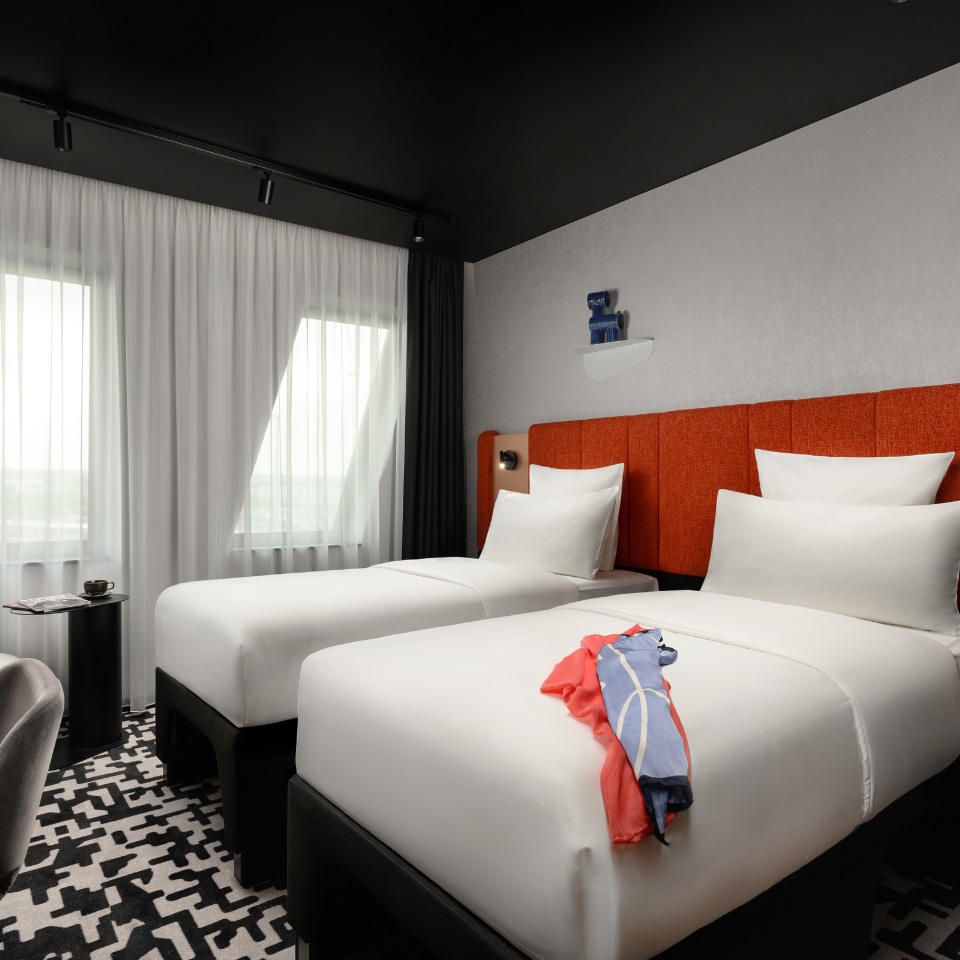 Modern hotel room with two neatly made single beds featuring orange headboards. A colorful shirt is draped over one bed. Large windows with sheer curtains allow natural light. A sleek table holds a cup and magazine, enhancing the room's contemporary, tidy ambiance.