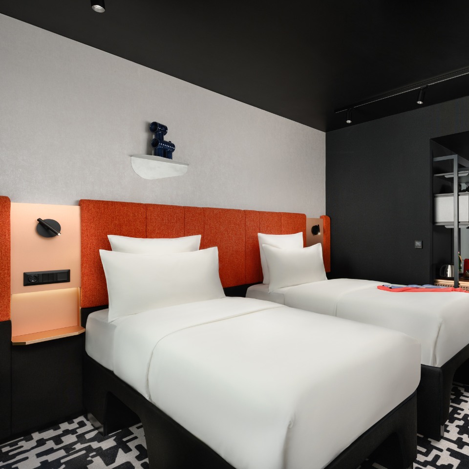 Modern hotel room with two neatly made single beds featuring orange headboards. The room has a black and white patterned carpet and a sleek black and gray color scheme.