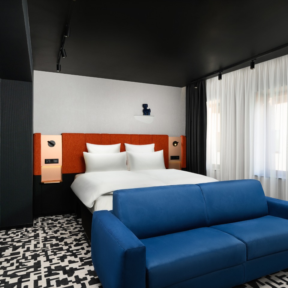 Modern hotel room with a black ceiling, bold orange headboard, white bedding, and a striking blue couch. A black-and-white patterned carpet adds contrast.