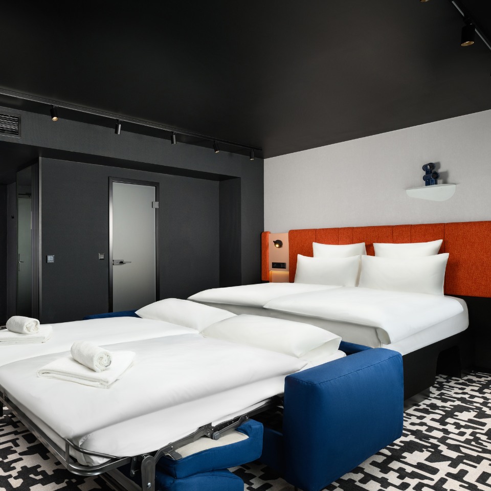 Modern hotel room with a double bed and blue pull-out sofa bed. The room features bold black, white, and orange tones, creating a sleek, stylish ambiance.