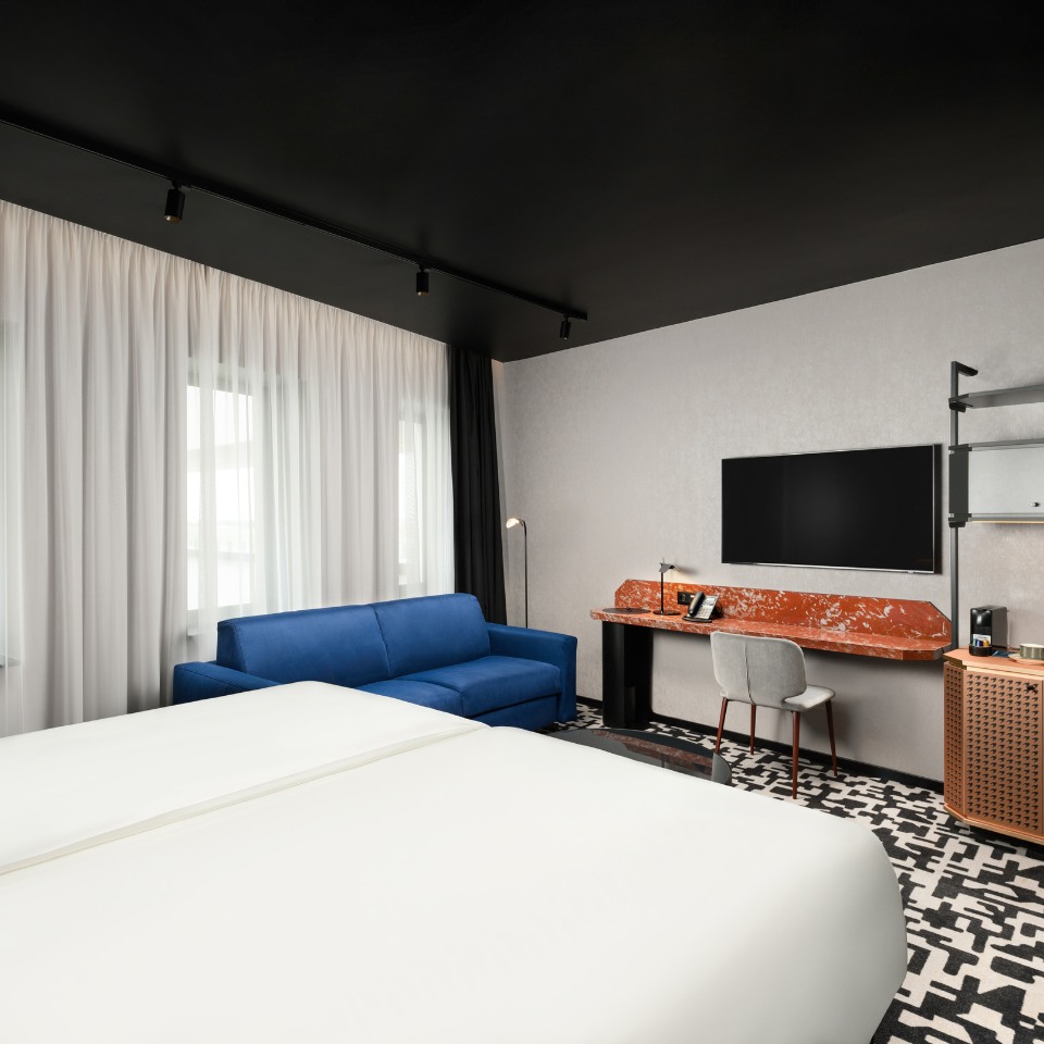 Modern hotel room with white twin beds, blue sofa, and patterned floor. Marble desk features a phone and TV, creating a chic, cozy atmosphere.
