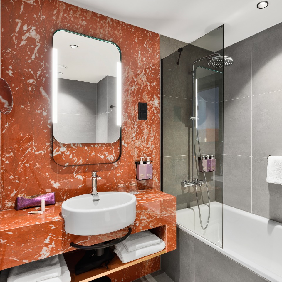 Modern bathroom with red marble countertop, circular white sink, and backlit mirror. Gray tiled walls, bathtub with glass screen, and towels neatly arranged. Elegant, clean design.