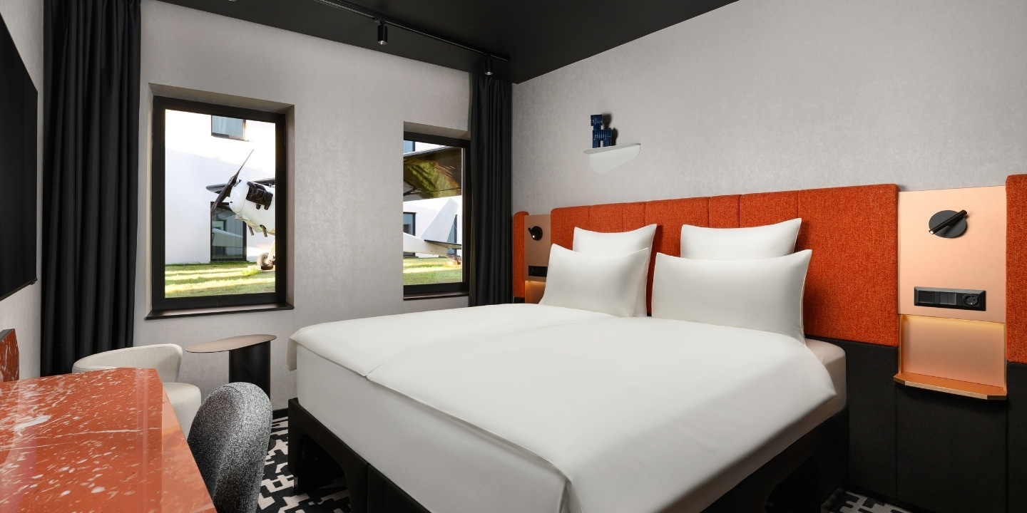 Modern hotel room with double bed featuring white linens and orange headboard. Large windows with dark curtains offer a view of green lawn outside.