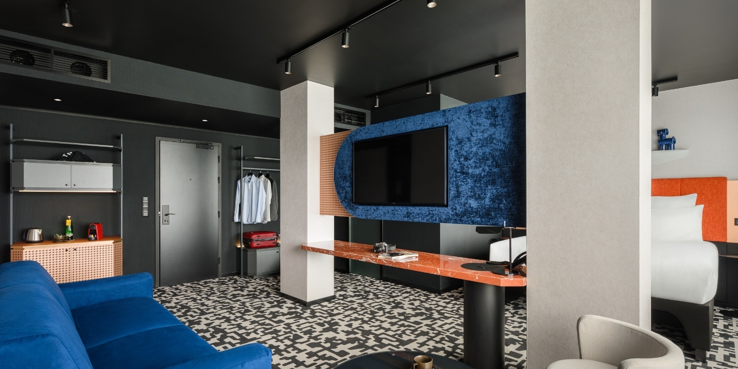 Modern hotel room with a blue couch, patterned carpet, and a large flat-screen TV on an orange and blue panel. Open closet with shirts and a suitcase.