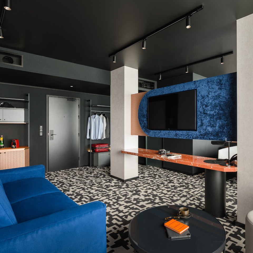 Modern hotel room with a bold design: blue sofa, red marble desk, large TV on blue velvet panel, patterned carpet, open closet, relaxed vibe.