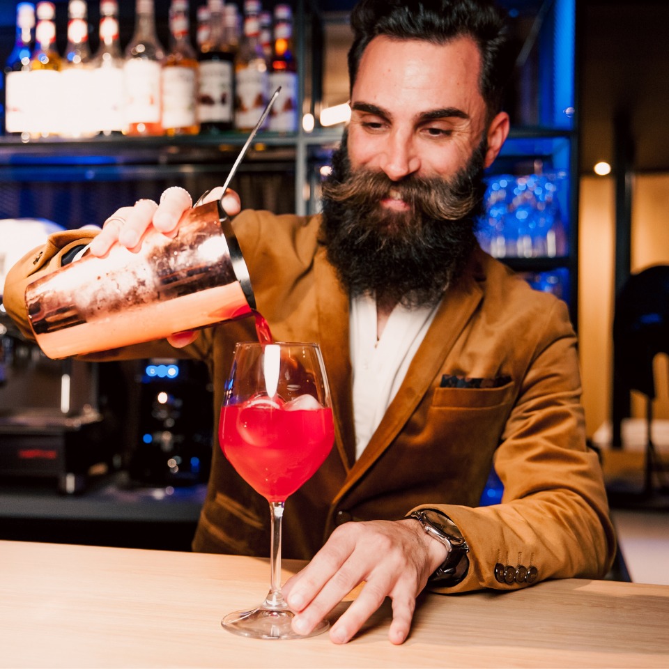 A bartender with a beard and a brown jacket is pouring a colorful drink from a shaker