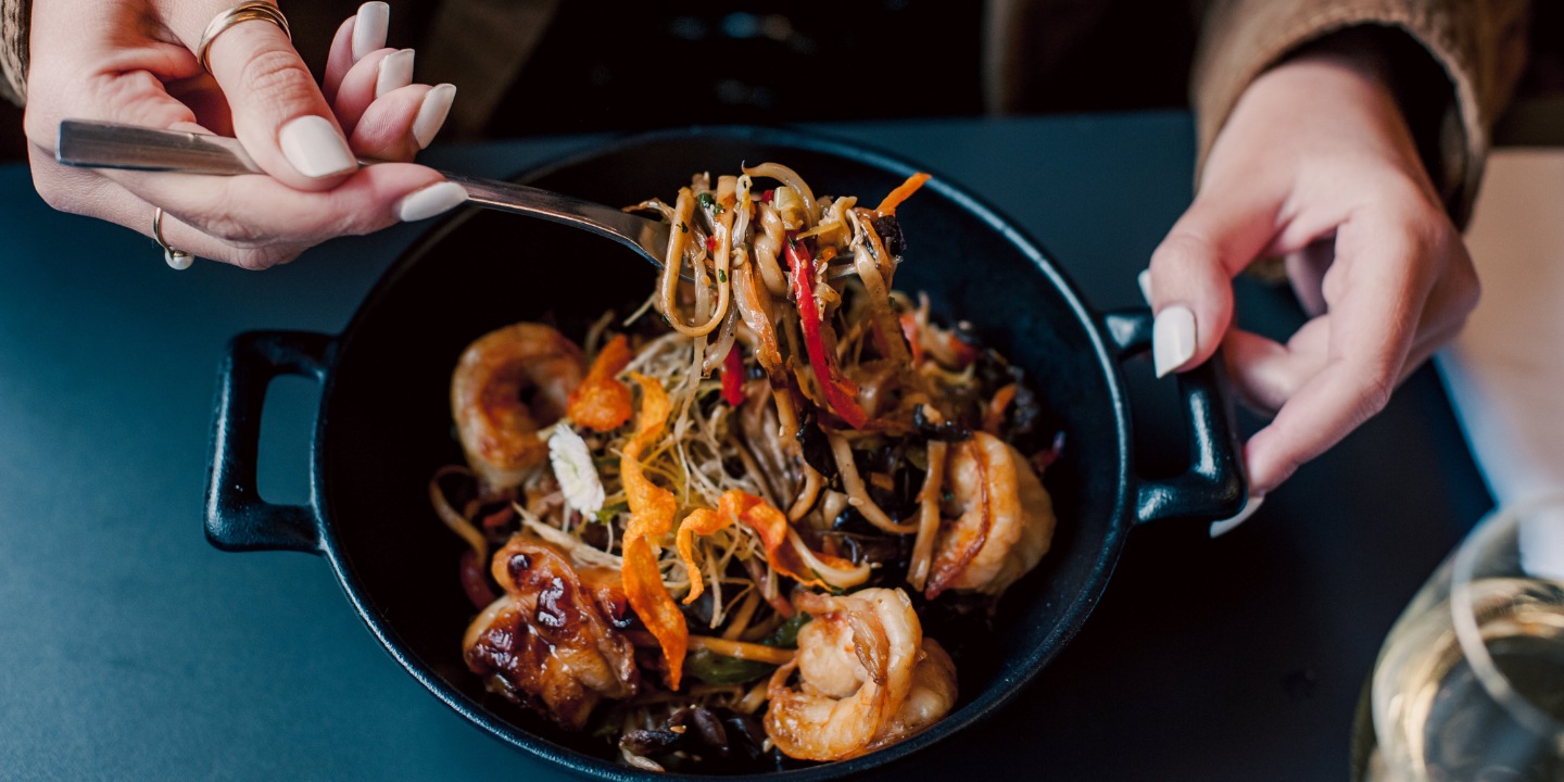 A person holds a fork filled with colorful stir-fried noodles and shrimp in a black bowl, set against a dark table.
