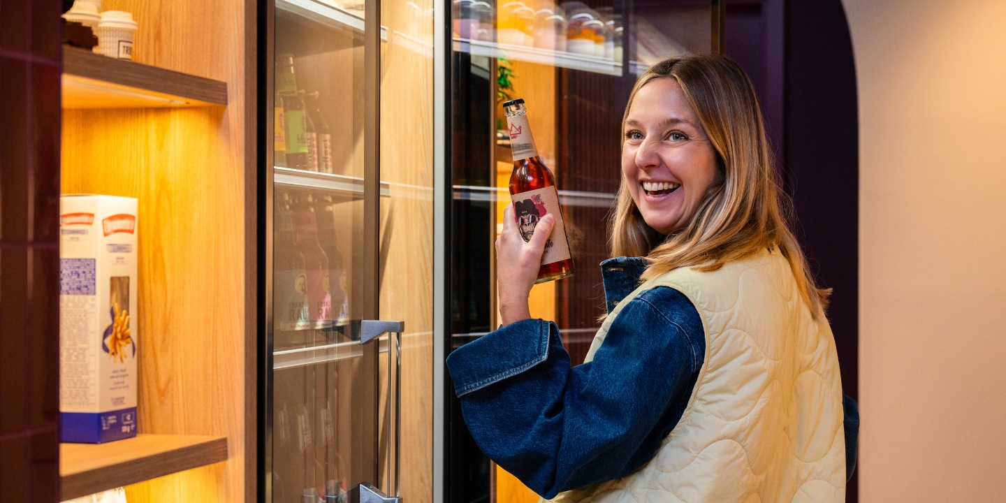 A person holding a bottle stands in front of a well-stocked, modern refrigerator filled with beverages and snacks.