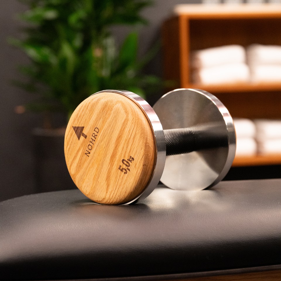 A 50 kg wooden dumbbell with a sleek metal handle rests on a black bench, set against a backdrop of towels and greenery.