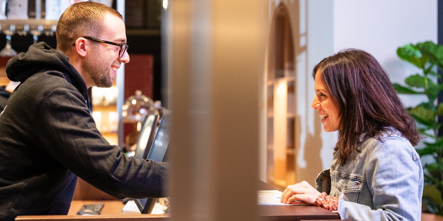 A person in a dark hoodie engages with another person at a welcoming reception desk, surrounded by warm wooden decor.