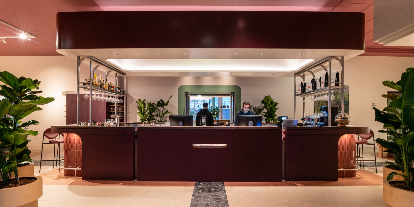 A modern bar with a sleek maroon counter, shelves of liquor, and potted plants, illuminated by soft lighting.