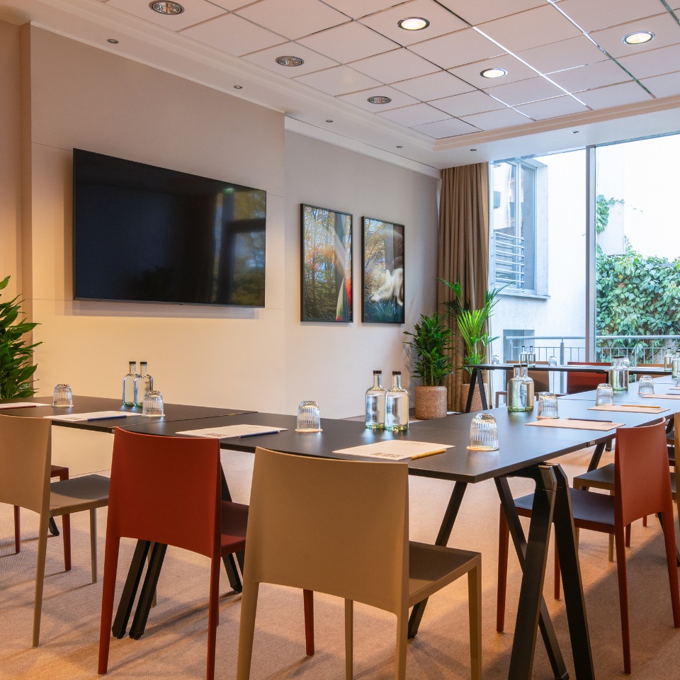 Modern conference room with a large screen, stylish chairs, glass water bottles, and greenery, bathed in natural light from a window.
