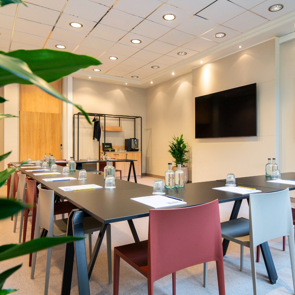 Modern conference room featuring a large table, colorful chairs, a wall TV, and refreshments on a side table.