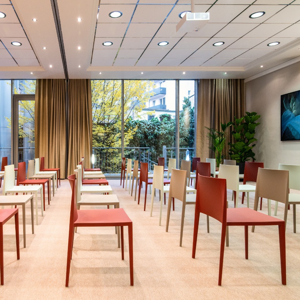 A modern meeting room featuring rows of colorful chairs and large windows showcasing greenery outside. Soft lighting enhances the atmosphere.