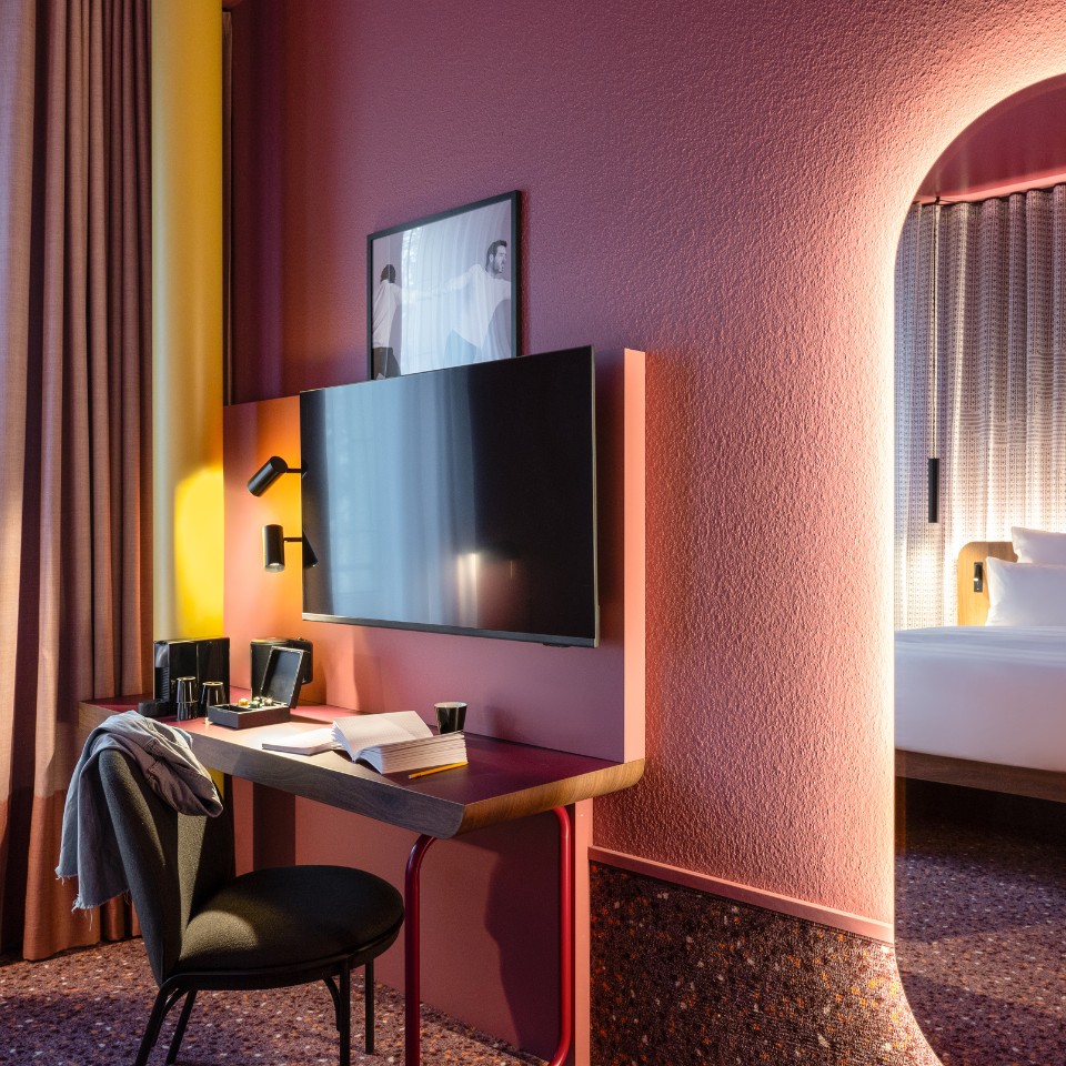 A hotel room corner featuring a stylish desk, wall-mounted TV, and vibrant colorful decor, with a glimpse of a bed beyond.